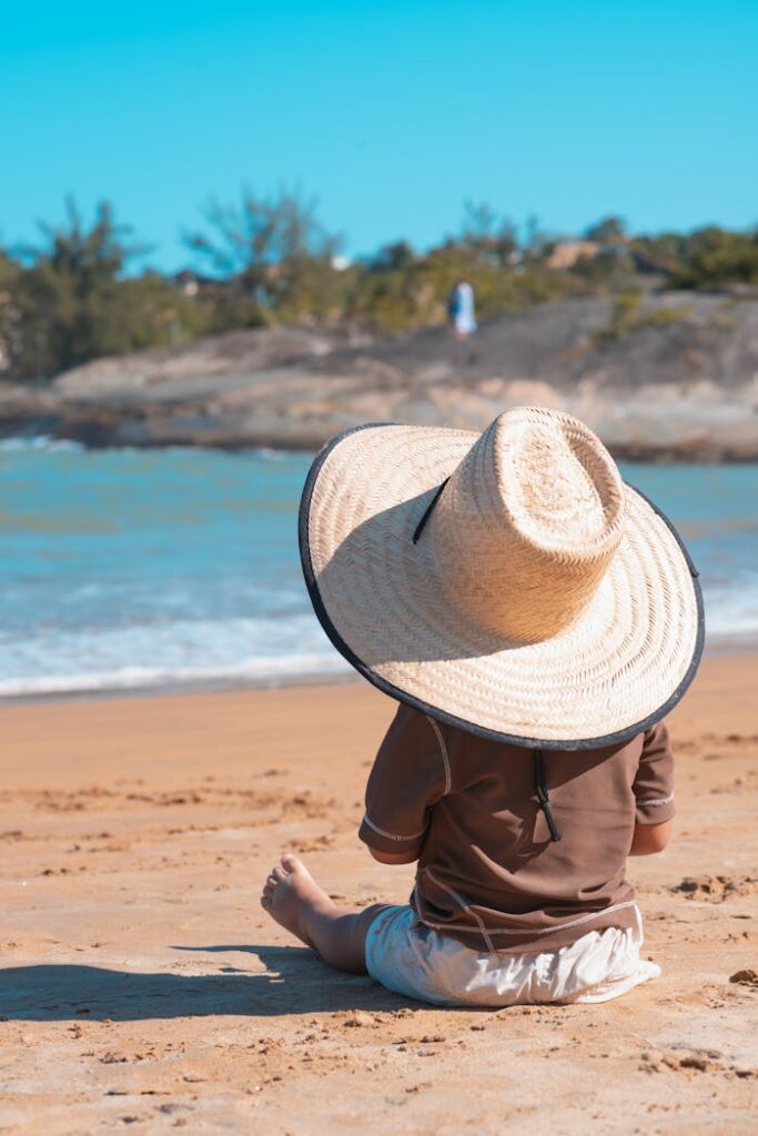 Home A young child wearing a large sunhat sits on a sandy beach by the ocean.